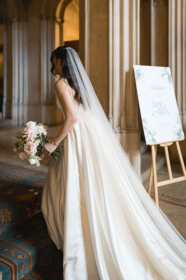 Allyn walking past her Ian & Allyn welcome sign, bouquet in hand, her pearl-scatter Coralie veil trailing to the floor over a satin gown.