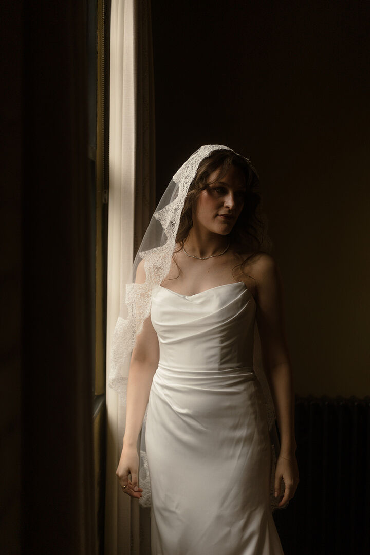 Rhiannon lit by a tall window, the eyelash lace edge of her Adela mantilla veil framing her face and shoulders.